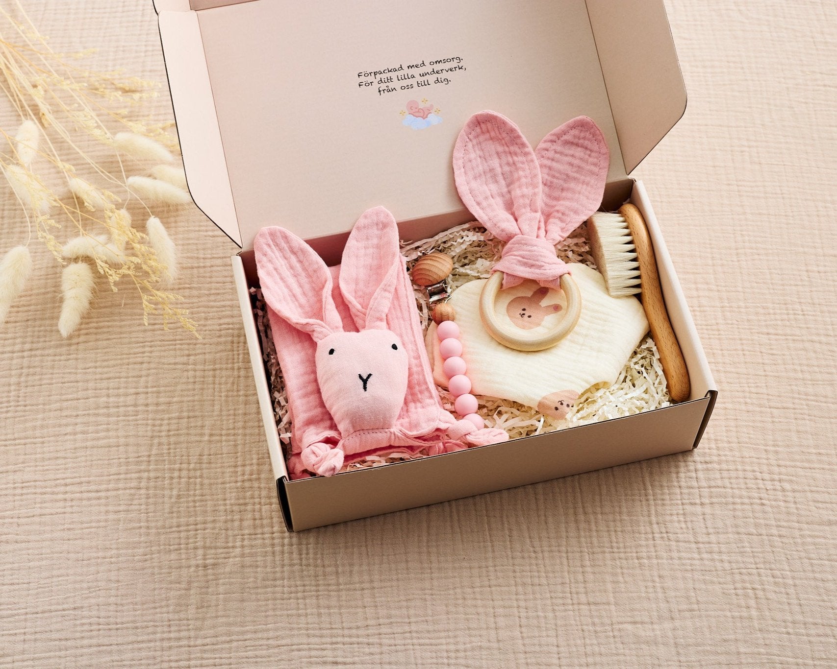 Rosa babyset i presentförpackning med snuttefilt formad som en kanin, bitring med tygöron, träborste, nappklipp med träpärlor och en beige muslinduk med björnmönster. Perfekt för nyfödda och babyshowers.